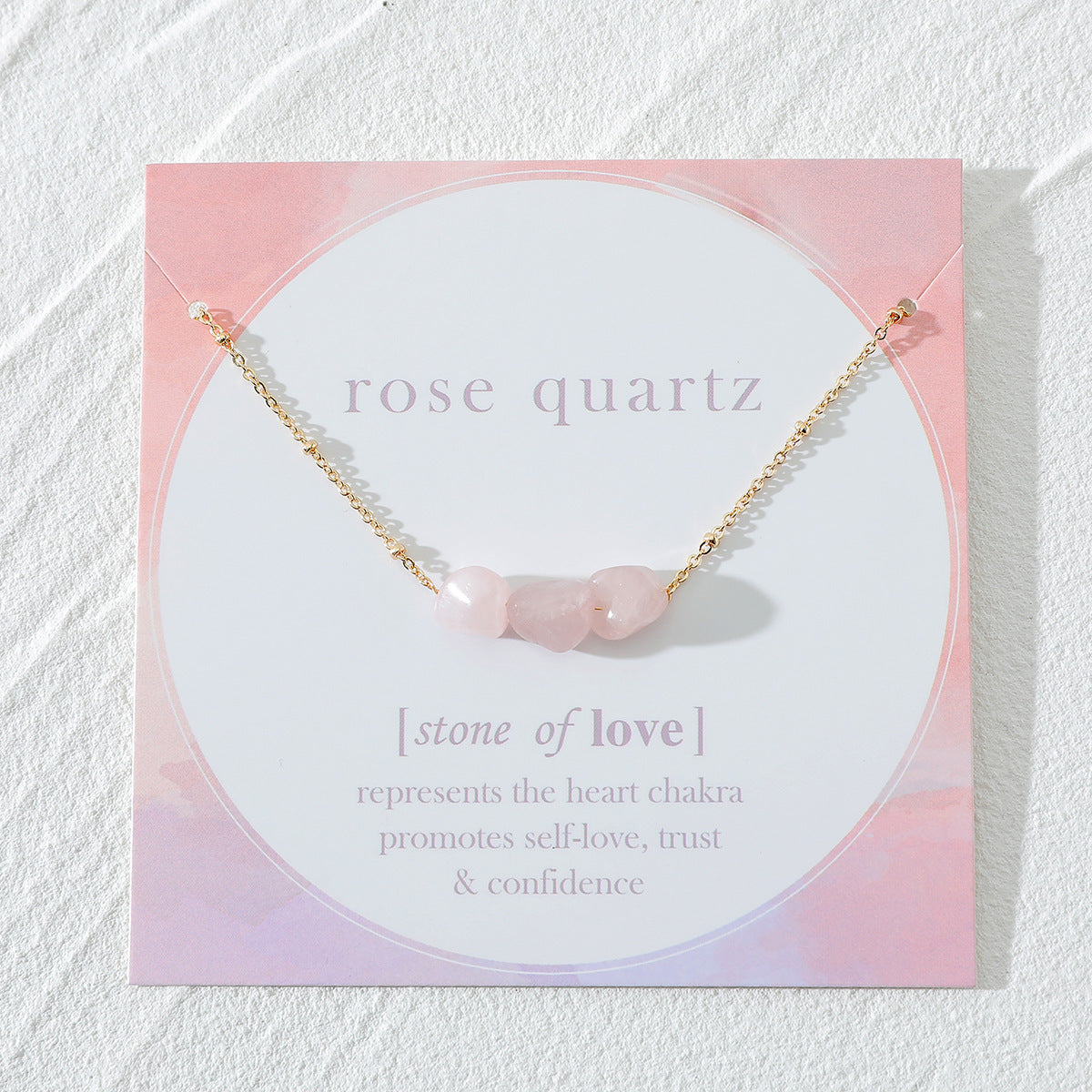 "Flat-lay view of a rose quartz necklace with raw gemstone beads on a gold chain, elegantly displayed on a card emphasizing rose quartz as the stone of love. The card highlights its connection to the heart chakra, promoting self-love, trust, and confidence. The soft pink hues complement the delicate design by Oyracha Jewelry."