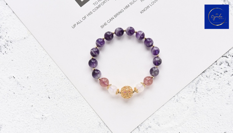 Top-down view of a handcrafted amethyst bracelet featuring polished purple amethyst beads and a decorative gold-tone bead centerpiece. The bracelet is beautifully arranged on a white textured surface, emphasizing its elegance and spiritual energy. Available now at Oyracha Jewelry.