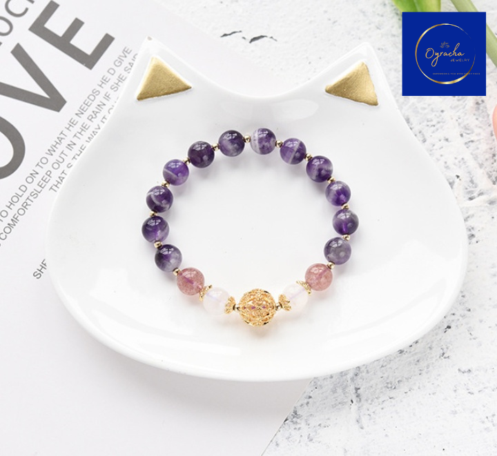 Top-down view of a handcrafted amethyst bracelet featuring polished purple amethyst beads and a decorative gold-tone bead centerpiece. The bracelet is beautifully arranged on a white textured surface, emphasizing its elegance and spiritual energy. Available now at Oyracha Jewelry.