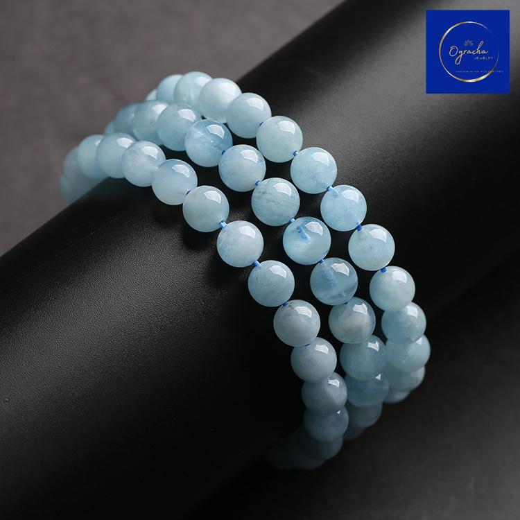 Side view of multiple aquamarine bead bracelets stacked on a black cylindrical surface, highlighting the smooth and polished finish of the beads. This tranquil aquamarine bracelet from Oyracha Jewelry radiates calm and serenity, perfect for enhancing one's sense of balance and peace.