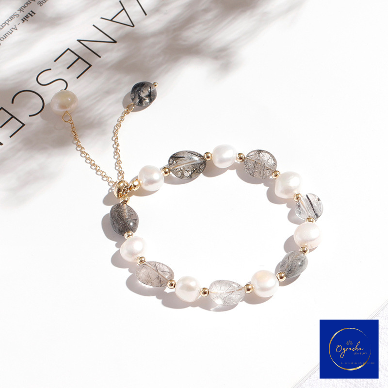 Top angled view of a moonstone and freshwater pearl bracelet by Oyracha Jewelry, featuring dark gray rutilated quartz stones paired with luminous pearls and gold bead accents. The bracelet is showcased on a minimal white background, highlighting its modern and sophisticated design.